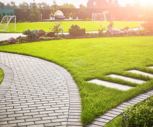 Landscaping of the garden. A tile path between green grass and a lawn with flowers in the sun. Soccer field in the background with copy space.; Shutterstock ID 1647173437; other: -; purchase_order: -; client: -; job: -