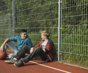 Two boys with basketball leanaing against fence