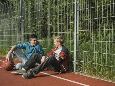 Two boys with basketball leanaing against fence